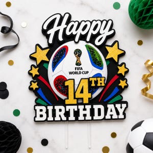 May include: A cake topper with the words "Happy 14th Birthday" in black and white. The design includes a soccer ball with the FIFA World Cup logo, gold stars, and colorful accents. The topper is designed for a birthday celebration.