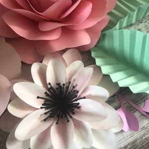 May include: A close-up of a paper flower craft. The flower is white with a black center and pink edges. The flower is surrounded by other paper flowers in pink and green.