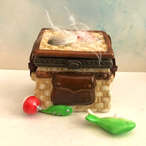 May include: A decorative trinket box shaped like a fishing tackle box. The box is tan with a brown lid and accents, and a brown leather-like pouch on the front. A red and white bobber, a green lure, and a green fish figurine are nearby.
