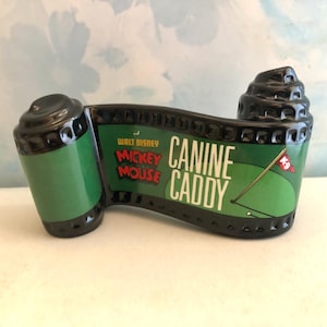 May include: A black and green ceramic filmstrip-shaped figurine with the words "Walt Disney Mickey Mouse Canine Caddy" printed on the front. The figurine has a golf-themed design with a flag and golf ball.