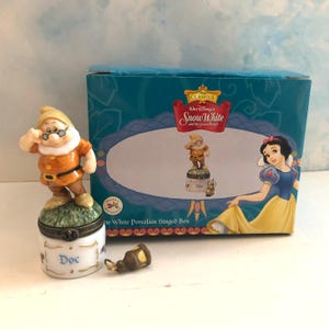 May include: A porcelain hinged box featuring a Doc figurine on the lid, with the name Doc printed on the side. The box is displayed next to its original teal packaging, which shows an illustration of Snow White and the Doc figurine.