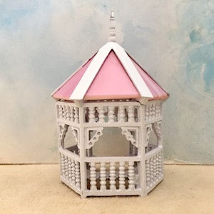 May include: A miniature white gazebo featuring a hexagonal structure with a pointed roof. The roof panels are painted a soft pastel pink, while the frame, railings, and decorative corner brackets are finished in a crisp, solid white paint.