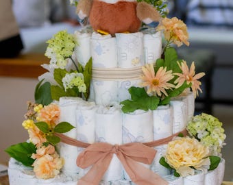May include: A three-tiered diaper cake decorated with flowers, ribbons, and a plush duck. The cake has a gold "Oh Baby" topper. The cake is decorated with light blue and white diapers.