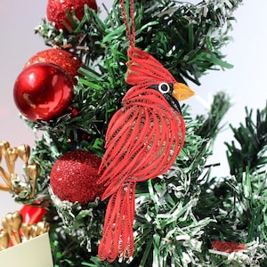 May include: A red cardinal bird ornament crafted from coiled paper strips, featuring a pointed crest, a black face mask, and a yellow beak. The bird is suspended by a thin red string and hangs among green pine needles and shiny red ornaments.