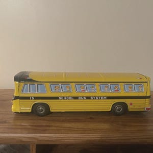 May include: A vintage yellow school bus toy with black accents and the text "SCHOOL BUS SYSTEM." The bus features windows with illustrations of children and the number "15" on the side. The toy is made of metal.