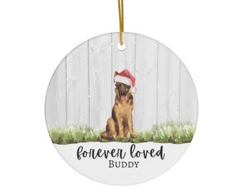 May include: A round ceramic ornament with a white woodgrain background and a German Shepherd dog wearing a red Santa hat. The dog is sitting on a patch of green grass with snow falling around it. The text "forever loved BUDDY" is printed below the dog.