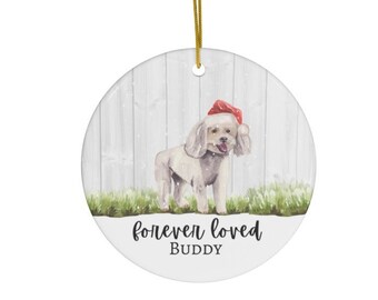 May include: White ceramic Christmas bauble featuring a watercolour illustration of a white dog wearing a red Santa hat. The bauble has the text "forever loved BUDDY" on the bottom.