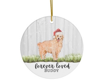 Könnte beinhalten: Ein weißer Keramik-Weihnachtsbaumschmuck mit einer Aquarellmalerei eines Golden Retrievers, der einen Weihnachtsmann-Hut trägt. Der Schmuck hat den Text "forever loved BUDDY" auf der Unterseite.