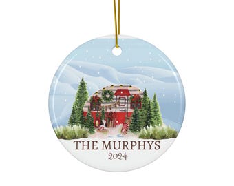 Op de afbeelding: Een ronde kerstbal van keramiek met een winterscène met een rood-wit gestreepte camper die versierd is voor de feestdagen. De kerstbal heeft een blauwe achtergrond met witte sneeuw en bomen. De tekst "THE MURPHYS 2024" staat onderaan gedrukt.