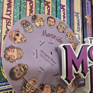 以下が含まれることがあります： 紫と白の「Monkey Island」のロゴが、キャラクターの顔が描かれたゲームホイールの前に立っています。ホイールには「Marca-Un-」と「Lucasfilm Games」の文字があります。ロゴの後ろには、いくつかのゲームボックスがあります。