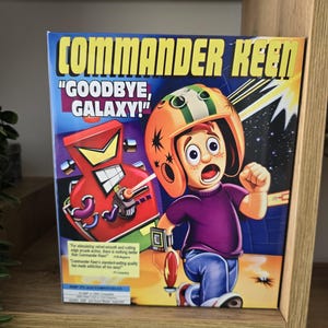 Könnte beinhalten: Eine Commander Keen Videospielbox mit dem Text "GOODBYE, GALAXY!". Das Cover zeigt einen Cartoon-Jungen mit Helm, einen roten Roboter und eine Weltraumszene. Die Box wird auf einem Holzregal ausgestellt.