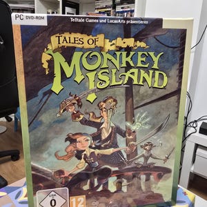 Puede incluir: Caja de juego para PC DVD-ROM de "Tales of Monkey Island". La ilustración presenta una temática pirata con personajes empuñando espadas y un mono. El título está en letras grandes y amarillas. La caja tiene una clasificación USK de 0 y 12.
