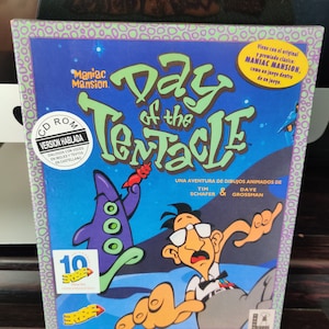 Può includere: Scatola CD-ROM del gioco "Day of the Tentacle" con un'illustrazione a cartoni animati. La scatola è prevalentemente blu con il titolo del gioco in grandi lettere verdi. L'artwork include un tentacolo viola e un personaggio sorpreso.