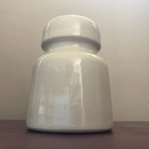 以下が含まれることがあります： 丸みを帯びた上部と幅広のベースを備えた白いセラミックボトル。 ボトルは滑らかで光沢のある仕上げとシンプルでクラシックなデザインです。 ボトルは暗い表面に置かれています。
