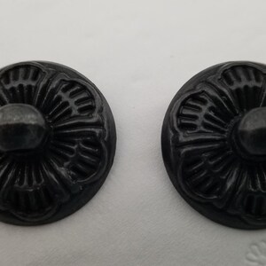 May include: Two black metal decorative buttons with a floral design. The buttons have a raised center and a detailed floral pattern around the edge.