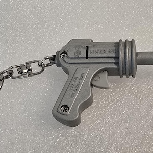 May include: A silver-colored toy gun keychain. The keychain features a small, detailed replica of a gun with a trigger, barrel, and handle. The gun is attached to a chain and a keyring. The text "TIMBERLAND" is visible on the side of the gun.