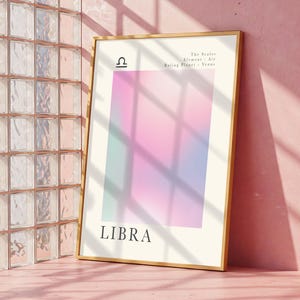 May include: A framed Libra zodiac sign art print. The artwork features a gradient of pink, purple, and blue colors. The Libra symbol and the word "LIBRA" are displayed in white text. The frame is gold.
