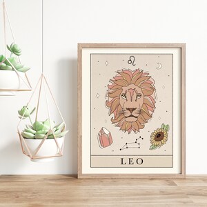 May include: A beige and brown illustrated print of the Leo zodiac sign. The print features a lion's head with a pink mane, a crystal, a sunflower, and the constellation Leo. The text "LEO" is printed at the bottom of the print.
