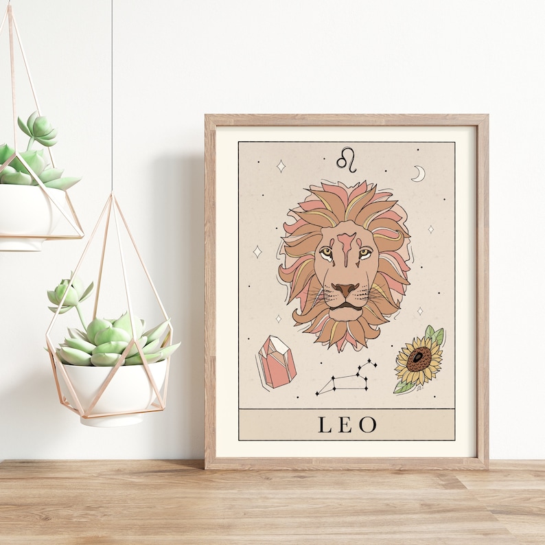 May include: A beige and brown illustrated print of the Leo zodiac sign. The print features a lion's head with a pink mane, a crystal, a sunflower, and the constellation Leo. The text "LEO" is printed at the bottom of the print.