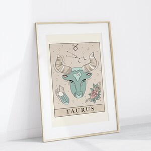 May include: A gold framed print with a beige background featuring a line drawing of a bull's head with a constellation of stars above it. The bull's head is teal blue and the constellation is brown. The print also features a crystal and two pink roses. The text "TAURUS" is printed below the bull's head.