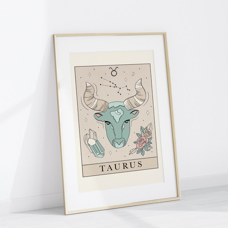 May include: A gold framed print with a beige background featuring a line drawing of a bull's head with a constellation of stars above it. The bull's head is teal blue and the constellation is brown. The print also features a crystal and two pink roses. The text "TAURUS" is printed below the bull's head.