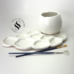 May include: A white ceramic paint palette with multiple circular wells and a matching white ceramic water cup. Two paintbrushes with dark blue handles and light-colored bristles rest across the palette. The logo Sally Schramm Pottery is visible.