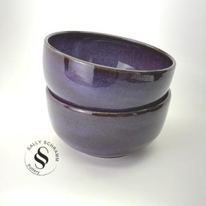 May include: Two stacked ceramic bowls with a deep purple, glossy glaze featuring subtle dark speckles. The bowls have a smooth, rounded shape. A circular logo in the bottom left corner displays the text Sally Schramm Pottery with a stylized S symbol.