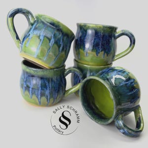 May include: Four ceramic mugs with a glossy glaze featuring a blend of deep blue and vibrant lime green colors. The glaze drips down the sides of the rounded vessels. A circular logo with the text Sally Schramm Pottery is visible below.