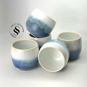 May include: Four ceramic cups featuring a white and blue ombre glaze with a drip pattern. The cups are arranged in a cluster. A circular logo with the text Sally Schramm Pottery and a stylized double S symbol is visible on the left side.