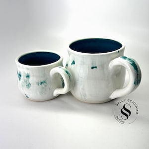 May include: Two white ceramic mugs with teal accents. The mugs have a dark teal interior and teal-colored handles. The larger mug has a wider base than the smaller mug. The mugs are handmade and have a unique, artistic design.