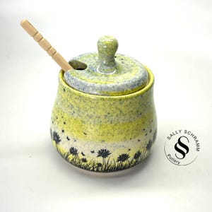 May include: Yellow and blue speckled ceramic honey pot with a wooden dipper. The pot has a black silhouette of dandelions and flying seeds. The bottom of the pot has a black circle with the text "Sally Schramm Pottery".