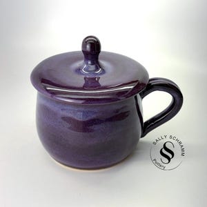 May include: A handmade, purple ceramic mug with a matching lid. The mug has a rounded shape, a curved handle, and a glossy finish. The lid has a small knob on top. The mug is suitable for hot beverages.