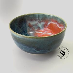 May include: A ceramic bowl featuring a deep blue and dark grey exterior glaze. The interior displays a vibrant blend of orange, red, and soft blue tones with white highlights. The text Sally Schramm Pottery is printed in black near the bottom right.