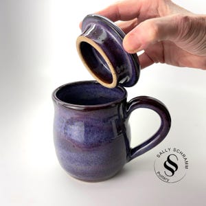 Puede incluir: Una taza de cerámica morada hecha a mano con una tapa a juego. La taza tiene una forma redondeada con un borde y asa marrón oscuro. La tapa se levanta, revelando el interior. La taza está marcada con el logotipo del artista.