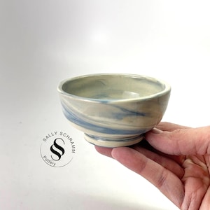 May include: A small, handmade ceramic bowl with a light blue and white marbled design. The bowl is held in a hand, showcasing its rounded shape and pedestal base. The bowl is likely used for serving small portions of food or as a decorative item.