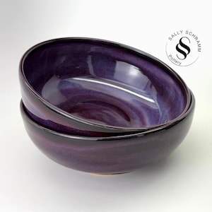 Puede incluir: Dos cuencos de cerámica apilados con un esmalte brillante de color púrpura intenso que se transforma en tonos violetas más claros. Las superficies interiores y exteriores tienen un acabado liso y reflectante. Se ve el logo Sally Schramm Pottery.