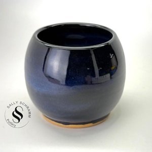 May include: A round ceramic pot with a glossy, deep navy blue glaze that transitions to a lighter blue hue. The base of the pot is unglazed, revealing a natural tan clay color. A circular logo with the text Sally Schramm Pottery is visible.