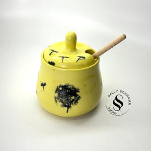 May include: Yellow ceramic honey pot with a black dandelion design and a wooden honey dipper. The pot has a lid with a small hole for the dipper. The pot is decorated with the text "Sally Schramm Pottery".