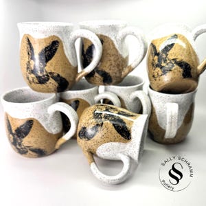 May include: A collection of handmade ceramic mugs. Each mug features a speckled white rim, a tan body with black abstract designs, and a white handle. The mugs are stacked and arranged, showcasing their unique, artistic style.