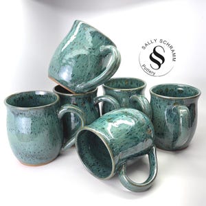 May include: A collection of five ceramic mugs with a glossy teal and dark green speckled glaze. Each mug features a curved handle and a rounded body. A circular logo with the text Sally Schramm Pottery is visible in the upper right corner.