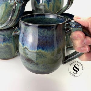 May include: A collection of ceramic mugs featuring a glossy glaze in shades of deep blue, navy, and moss green. Each mug has a rounded body and a sturdy handle. A circular logo with the text Sally Schramm Pottery is visible in the corner.