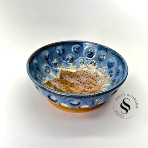 May include: A blue and brown ceramic bowl with a textured surface and a raised rim. The bowl has a speckled glaze and a small logo that says "Sally Schramm Pottery".