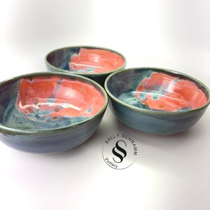 Puede incluir: Tres cuencos redondos de cerámica con un esmalte brillante que mezcla tonos azul profundo y naranja coral vibrante. Las superficies interiores muestran un efecto jaspeado. Un logotipo circular con el texto Sally Schramm Pottery es visible.