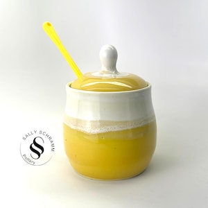 May include: A handmade ceramic sugar bowl with a lid and a yellow spoon. The bowl is yellow with a white band and a white knob on the lid. The spoon is yellow. The bowl is labeled with "SALLY SCHRAMM Pottery".