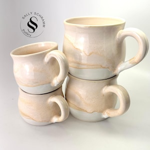 May include: A set of three ceramic mugs with a speckled, matte finish. The mugs are in shades of cream, light gray, and dark gray. Each mug has a simple, minimalist design and a rounded handle.