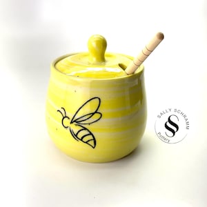 May include: A yellow ceramic honey pot with a lid and a wooden honey dipper. The pot features a black bee illustration and horizontal stripes. The logo "SALLY SCHRAMM Pottery" is on the right.