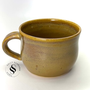 Puede incluir: Una taza de cerámica marrón y amarilla con forma redondeada y asa. La taza tiene un esmalte moteado y está hecha a mano.