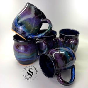 May include: A collection of four ceramic mugs featuring a glossy, deep purple and blue glaze with swirling iridescent highlights. Each mug has a rounded body and a curved handle. The text Sally Schramm Pottery is printed in black.