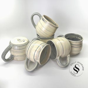 May include: A collection of handmade ceramic mugs. The mugs are off-white with gray and blue striped patterns and large gray handles. The mugs are stacked and arranged on a white surface. The logo "SALLY SCHRAMM Pottery" is visible.