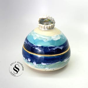 May include: A ceramic jar with a seashell-shaped lid. The jar is painted with horizontal bands of blue, turquoise, and white, resembling ocean waves. The lid is a detailed seashell. The jar is a unique decorative item.